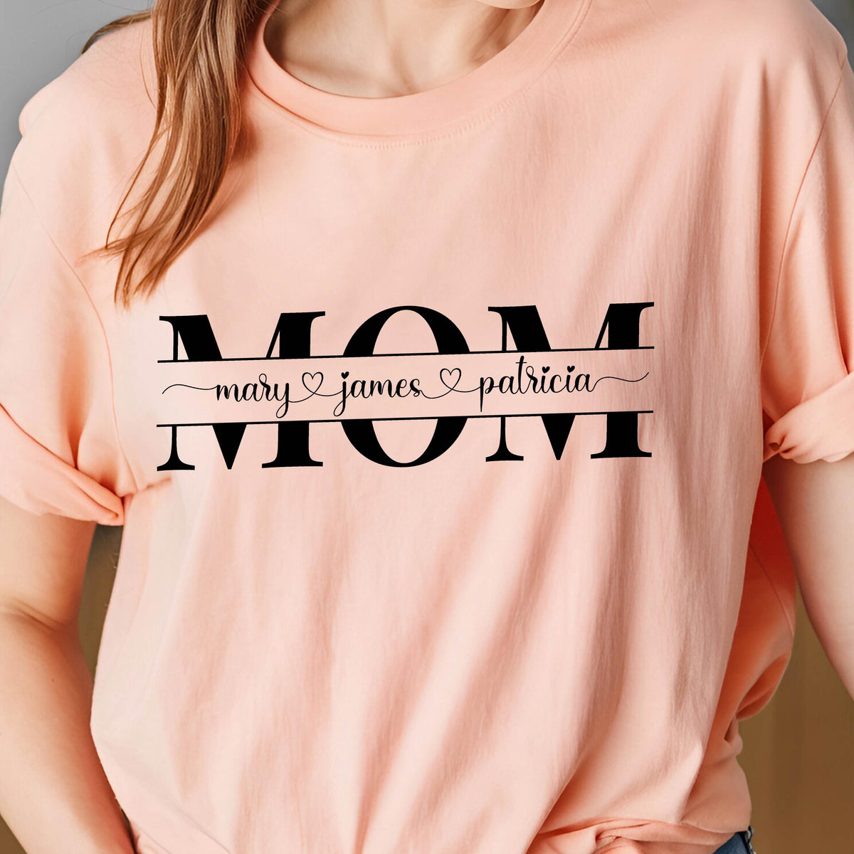 May include: A pink t-shirt with the word "MOM" in black letters. The word "MOM" is separated by a black line with the text "mary ♥ james ♥ patricia" written below it.