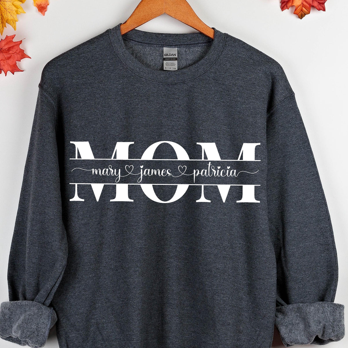 May include: Dark gray sweatshirt with the word "MOM" in white letters. Below the word "MOM" is a banner with the names "mary", "james", and "patricia" separated by hearts.