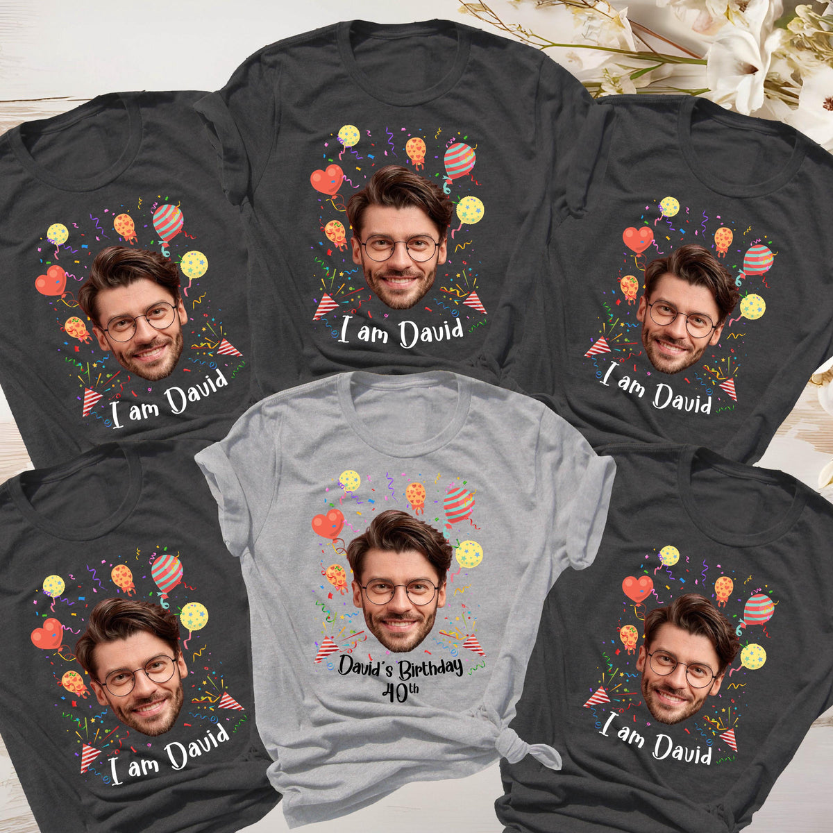 May include: Four dark gray t-shirts and one light gray t-shirt featuring a printed graphic of a man with glasses, surrounded by colorful balloons and confetti. The dark shirts display the text I am David, while the light shirt says David's Birthday 40th.