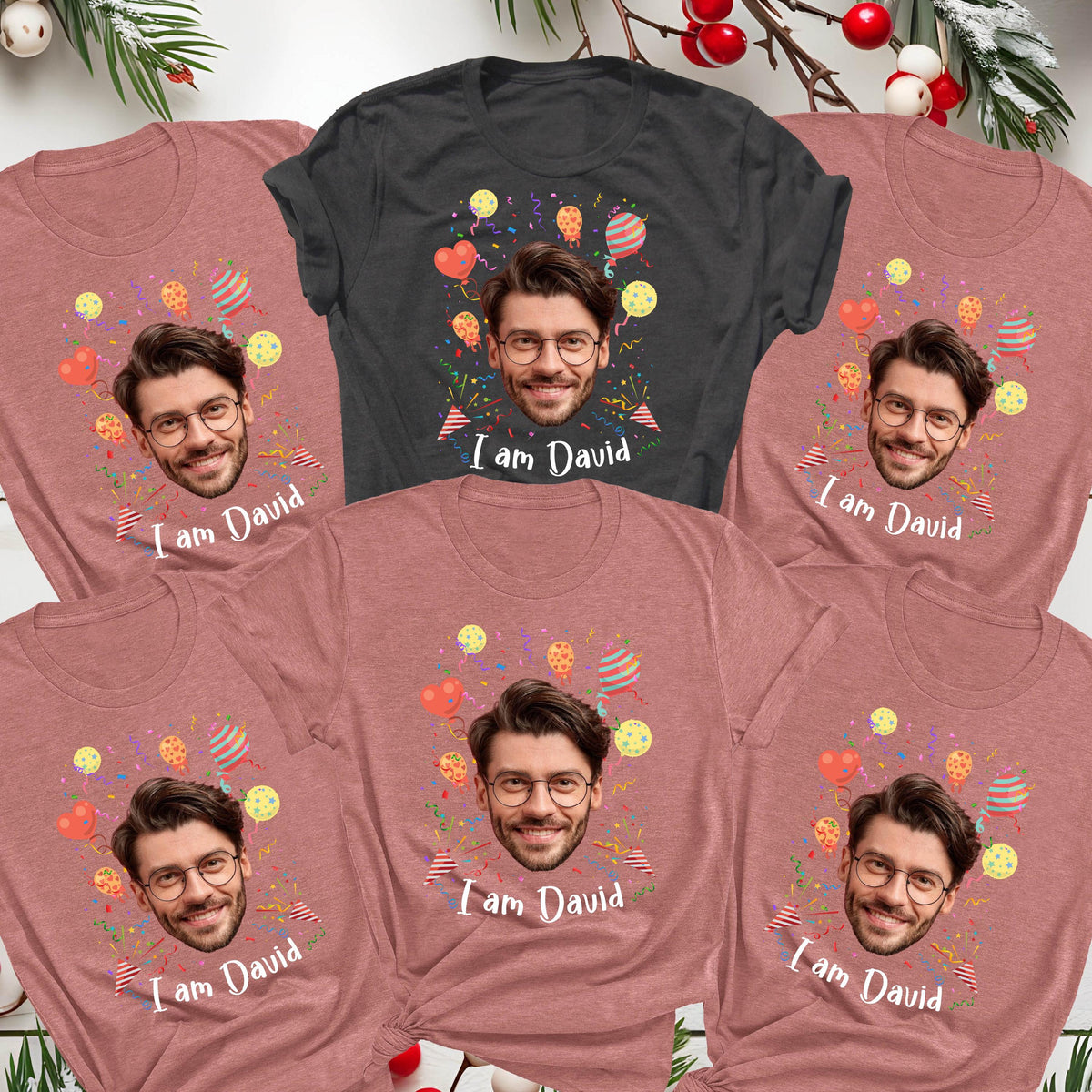 May include: Five short-sleeved t-shirts, one charcoal gray and four heather mauve, each featuring a centered photographic print of a man with glasses, surrounded by colorful balloons and confetti, with the text I am David printed below.