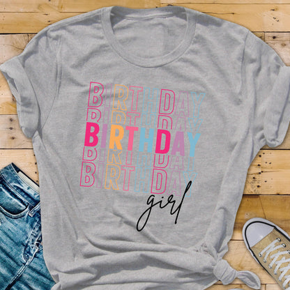 May include: A gray t-shirt with the word "BIRTHDAY" stacked vertically in pink, orange, and blue letters. The word "girl" is written in cursive below the stacked text. The shirt is folded at the bottom.