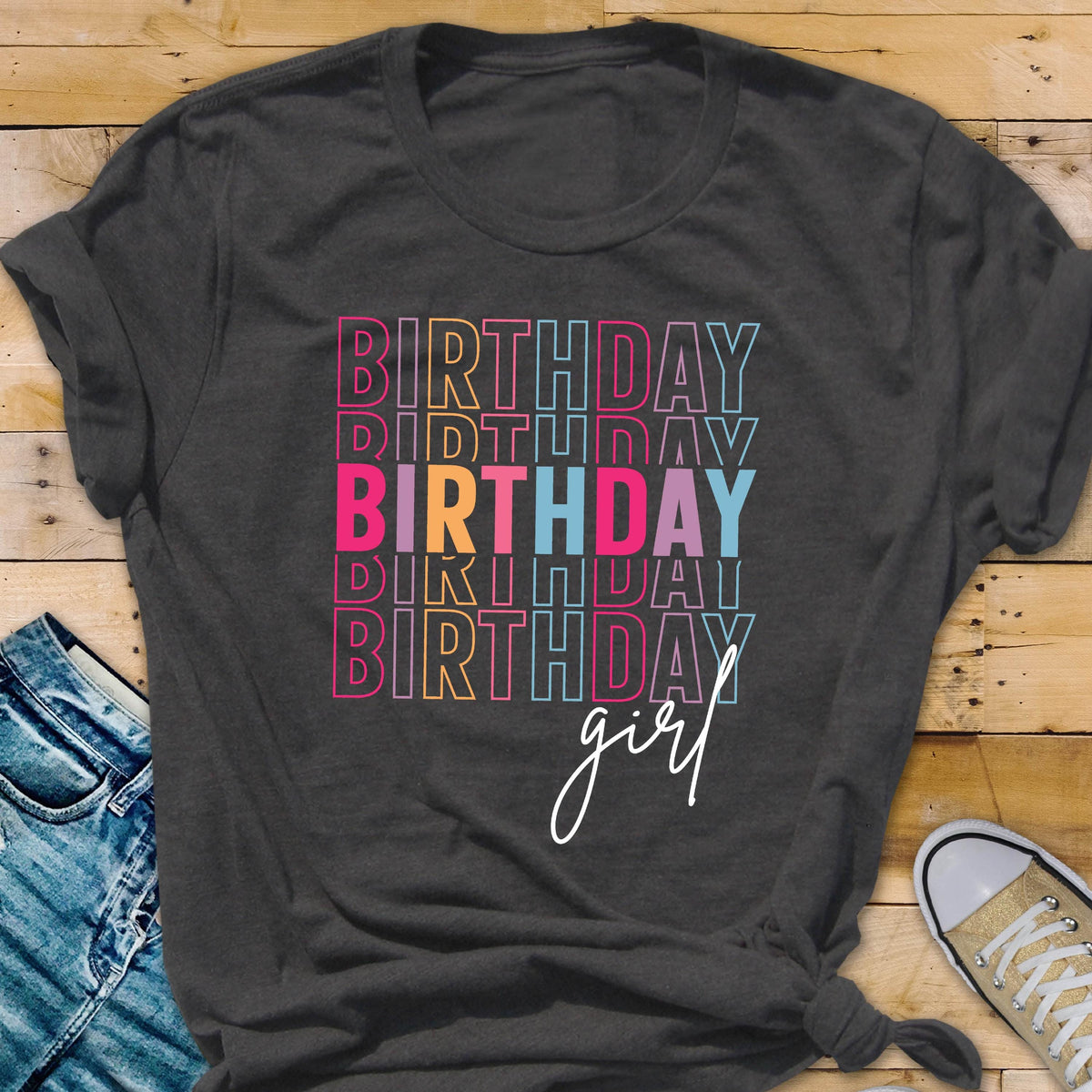 May include: Dark gray t-shirt with the word "BIRTHDAY" stacked in a colorful, retro font. The word "girl" is written in a cursive font below the stacked words. The shirt is a casual style.