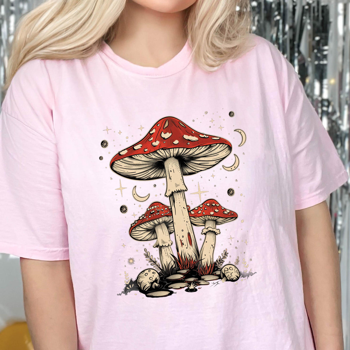 May include: Pink t-shirt with a graphic of three mushrooms with red caps and white stems. The mushrooms are surrounded by stars and crescent moons.