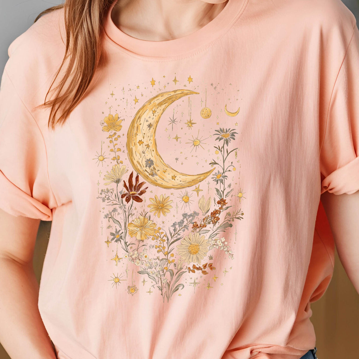 May include: A light pink t-shirt featuring a crescent moon and floral design. The moon is gold with a textured appearance, surrounded by yellow, white, and brown flowers, stars, and celestial elements. The shirt has rolled-up sleeves.