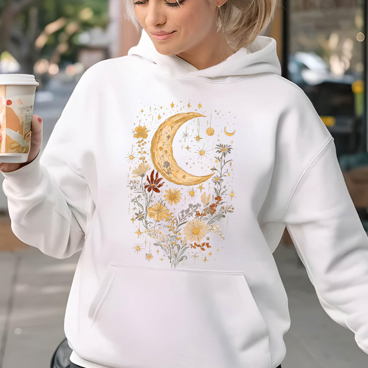 May include: A white hooded sweatshirt featuring a central graphic print of a large yellow crescent moon surrounded by various yellow and orange flowers, leaves, and small star shapes. The hoodie includes a front kangaroo pocket and long sleeves.