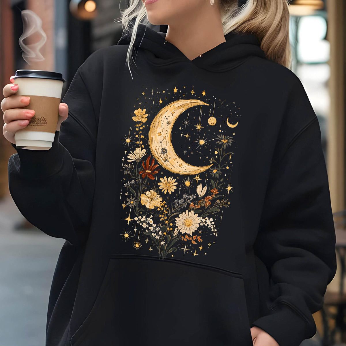 May include: A black hooded sweatshirt featuring a graphic print on the front. The design displays a large yellow crescent moon surrounded by various white and yellow flowers, small stars, and celestial elements arranged in a vertical composition.
