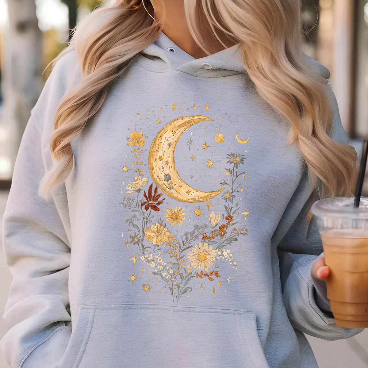 May include: A light blue hooded sweatshirt featuring a central graphic print of a large golden crescent moon surrounded by various yellow and orange wildflowers, small stars, and celestial elements, with a front kangaroo pocket and long sleeves.