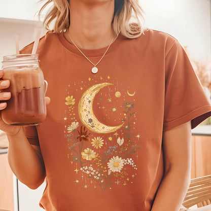 May include: A rust-colored t-shirt with a crescent moon and floral design. The moon is surrounded by small stars and flowers in shades of yellow, orange, and white.