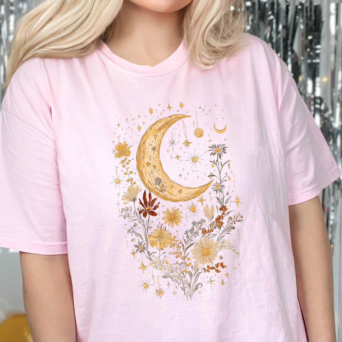May include: A pink t-shirt with a crescent moon and floral design. The moon is gold and surrounded by small stars and flowers in shades of yellow, orange, and brown.