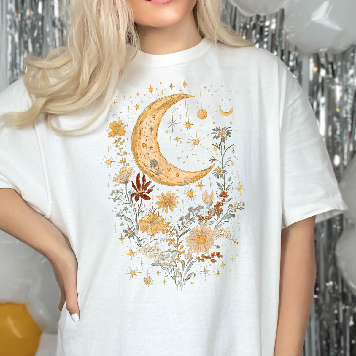 May include: White t-shirt with a graphic print of a crescent moon and flowers in shades of yellow, orange, and brown. The design is surrounded by small stars.