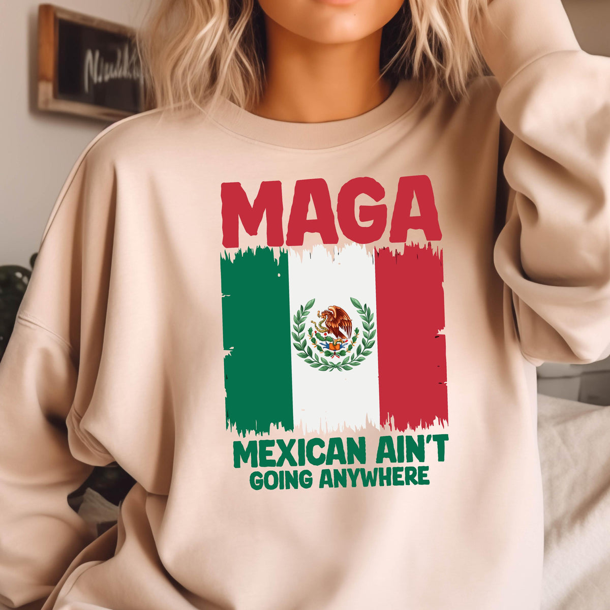 May include: A beige sweatshirt with the text "Maga Mexican Ain't Going Anywhere" printed on it. The text is in red, white, and green, and there is a stylized image of the Mexican flag behind the text.