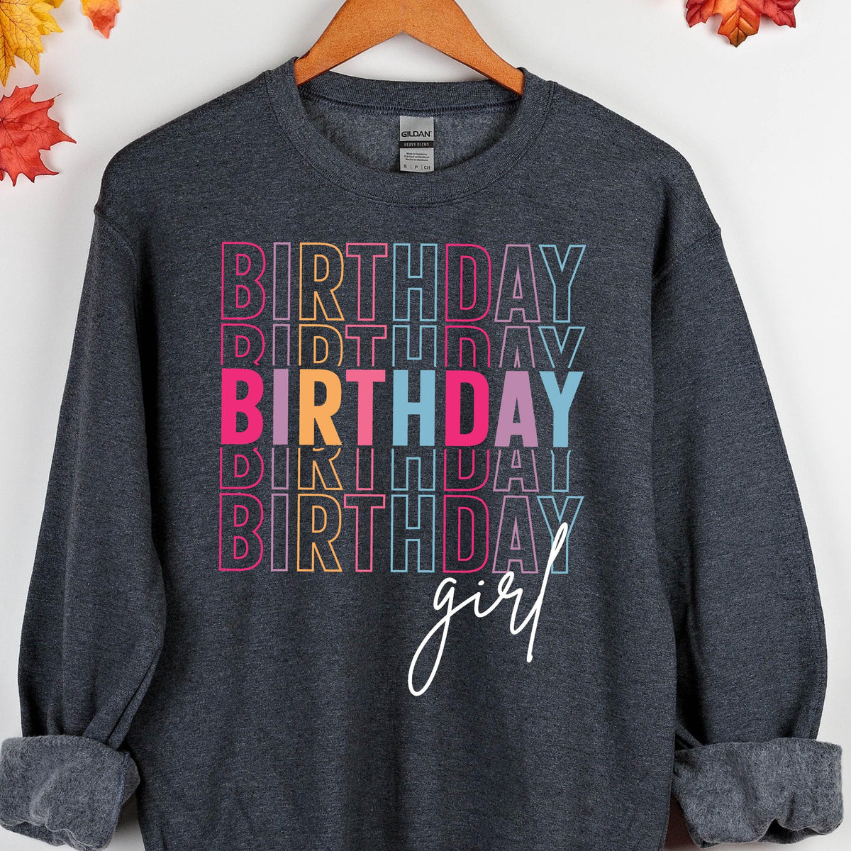 May include: A dark gray sweatshirt with the text "Birthday girl" in rainbow colors. The text is stacked vertically and the word "girl" is written in white cursive.