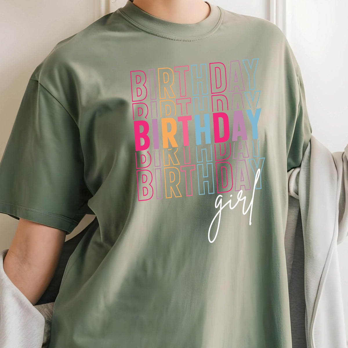 May include: A green t-shirt with the word "Birthday" repeated in rainbow colors and the word "girl" written in white script.