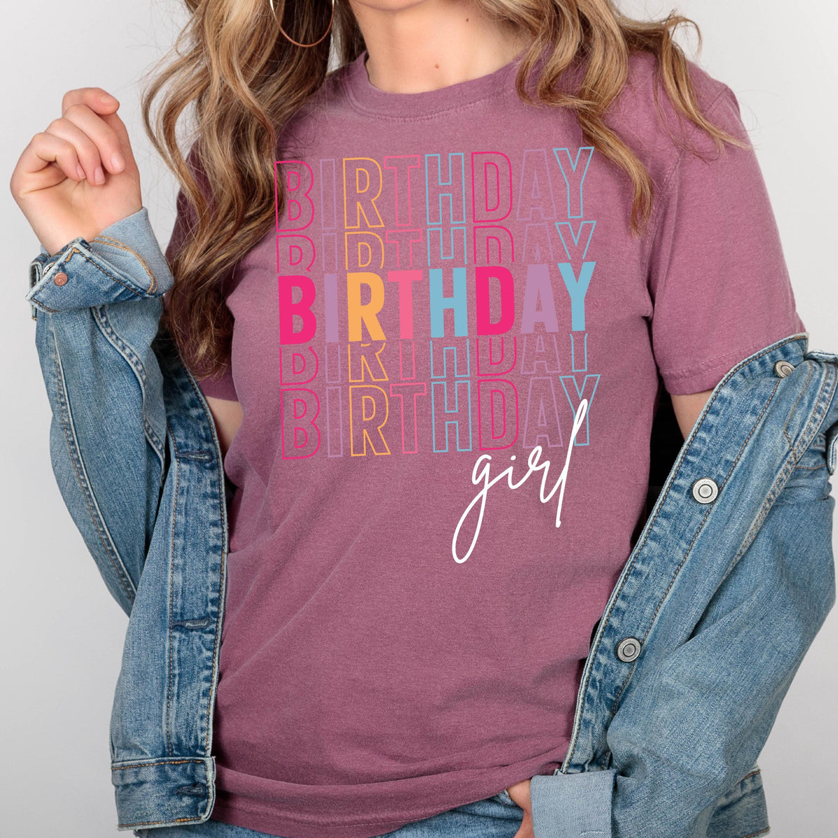May include: A mauve colored t-shirt with the text "BIRTHDAY girl" in rainbow colors. The text is stacked and repeated multiple times.