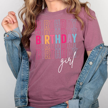 May include: A mauve colored t-shirt with the text "BIRTHDAY girl" in rainbow colors. The text is stacked and repeated multiple times.