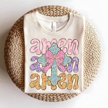 May include: A beige t-shirt with the word "amen" repeated three times in a colorful, floral design. The word "amen" is in a large, bold font and is surrounded by a floral pattern. A cross with a pink bow is in the center of the design.