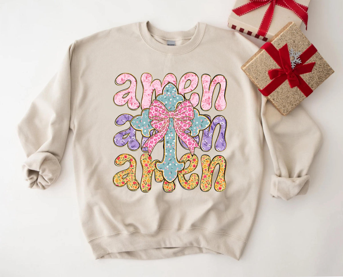 May include: A beige sweatshirt with the word "amen" written three times in a colorful, floral font. A cross with a bow is in the center of the design.