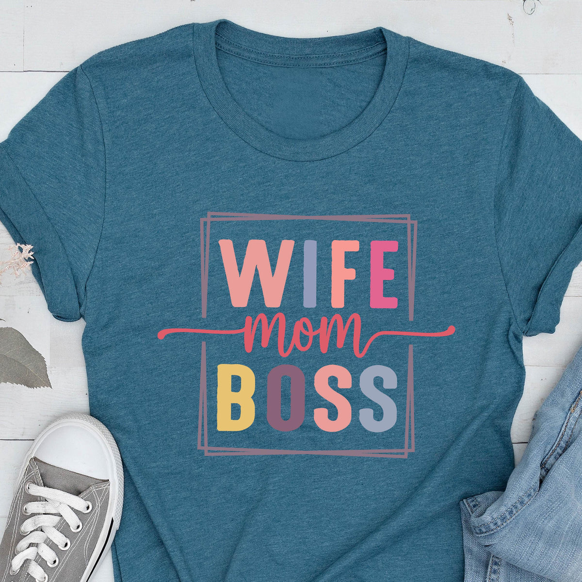 May include: Teal-colored t-shirt with the words "WIFE mom BOSS" in a colorful, rectangular design. The text is in various colors, including pink, orange, yellow, and blue. A gray canvas shoe is visible in the lower left corner.