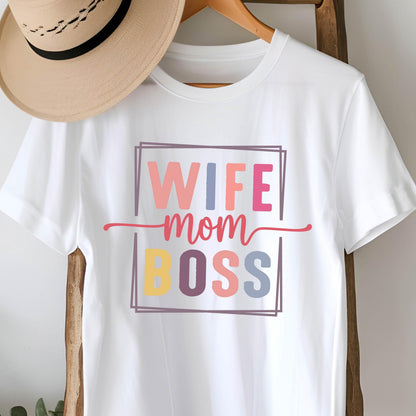 May include: White t-shirt with the words "WIFE mom BOSS" in a colorful, rectangular design. The text is in various colors, including pink, blue, yellow, and purple. A straw hat hangs on a wooden hanger above the shirt.