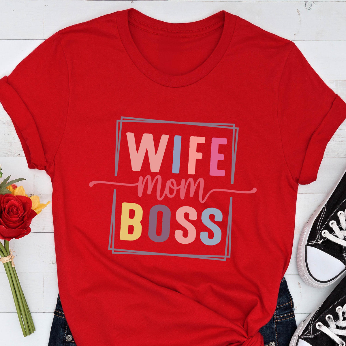 May include: A red t-shirt with the words "WIFE mom BOSS" in a colorful, rectangular design. The shirt is made of a soft material and has rolled-up sleeves. Black sneakers are visible in the bottom right corner.