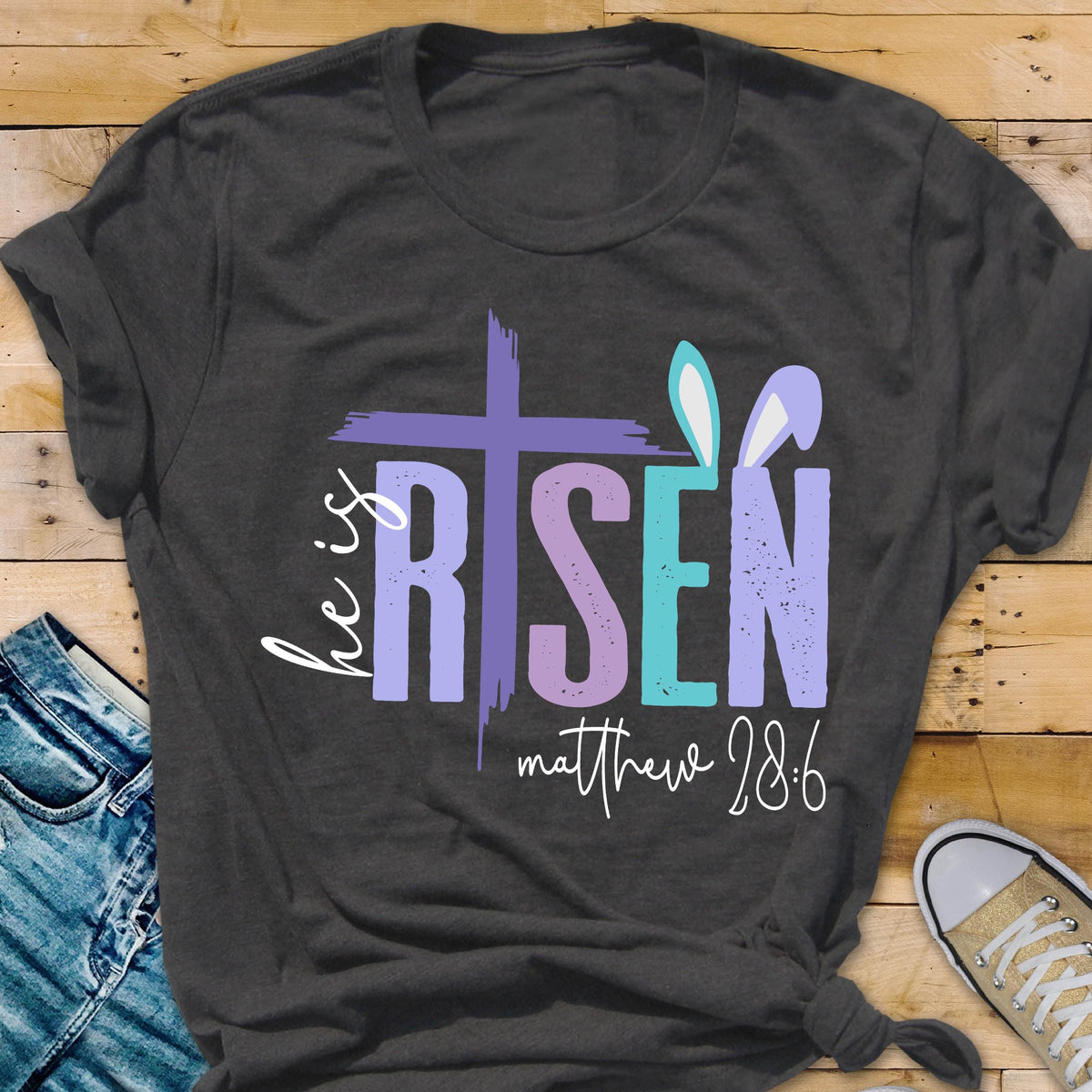 May include: A dark gray t-shirt with a colorful graphic design that says "He is Risen" with a cross and bunny ears. The text "Matthew 28:6" is below the graphic.