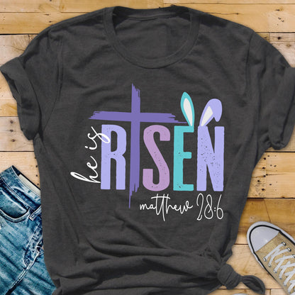 May include: A dark gray t-shirt with a colorful graphic design that says "He is Risen" with a cross and bunny ears. The text "Matthew 28:6" is below the graphic.