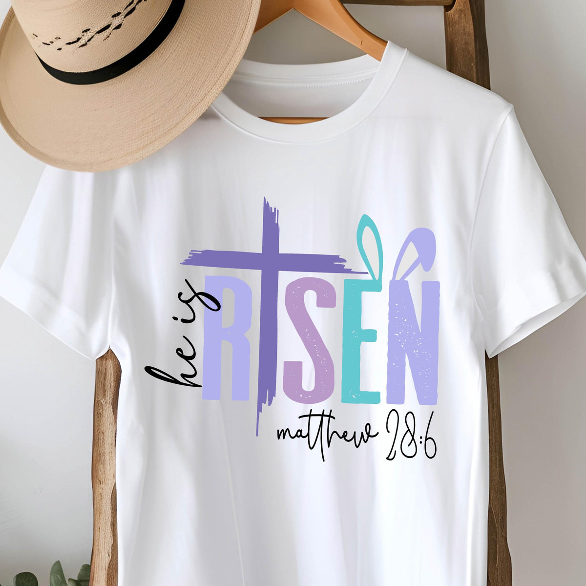 May include: White t-shirt with a colorful graphic design that says "He is Risen" with a cross and a bunny ear design. The text "Matthew 28:6" is also included.