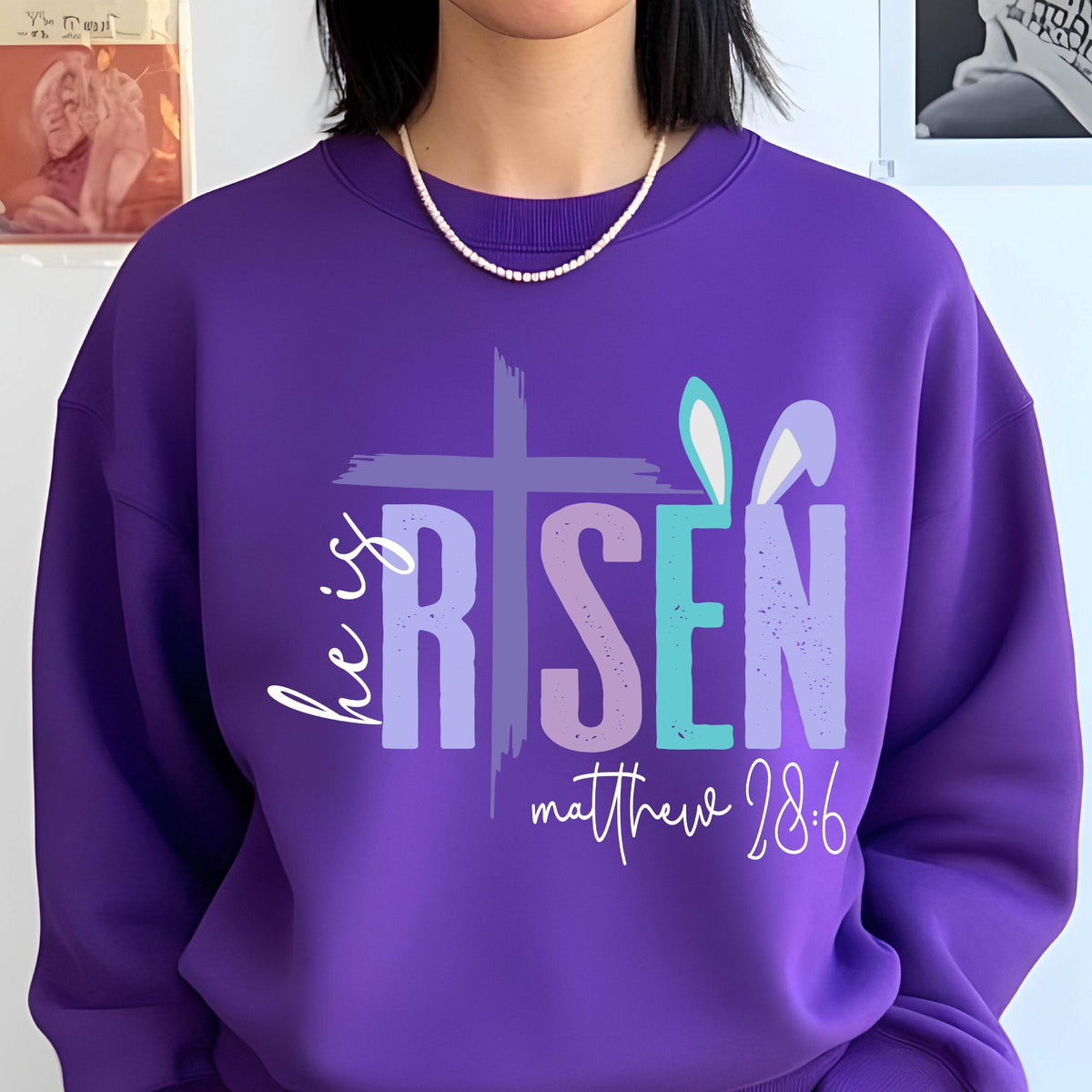 May include: Purple sweatshirt with a white cross and the words "He is Risen" in colorful letters. The text "Matthew 28:6" is below the words. There are bunny ears on the right side of the design.