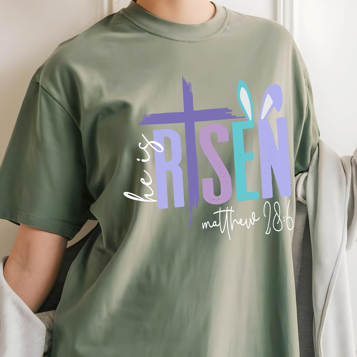 May include: Green t-shirt with a colorful graphic that says "He is Risen" with a cross and bunny ears. The text "Matthew 28:6" is below the graphic.