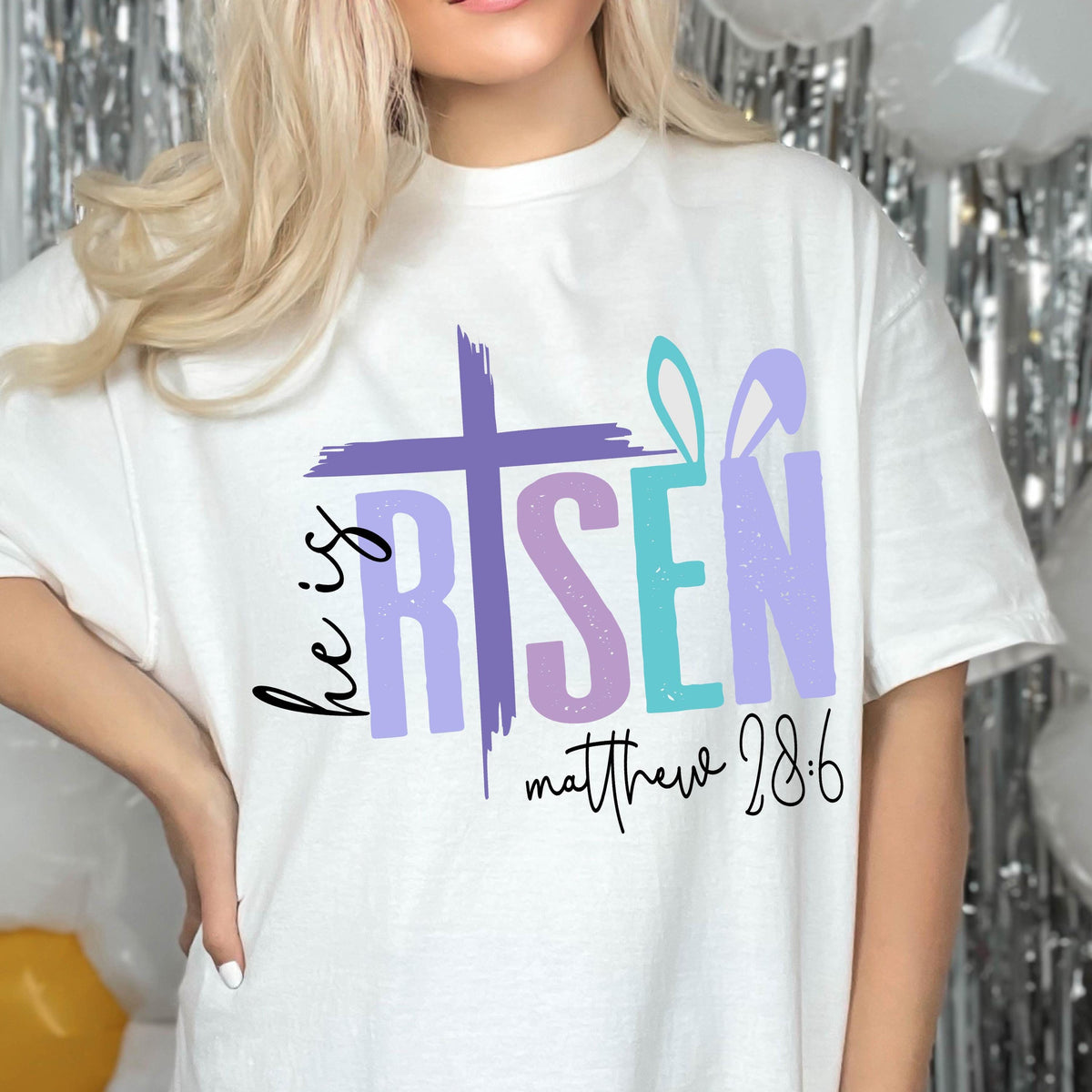 May include: White t-shirt with a colorful graphic design that says "He is Risen" with a cross and bunny ears. The text "Matthew 28:6" is below the graphic.