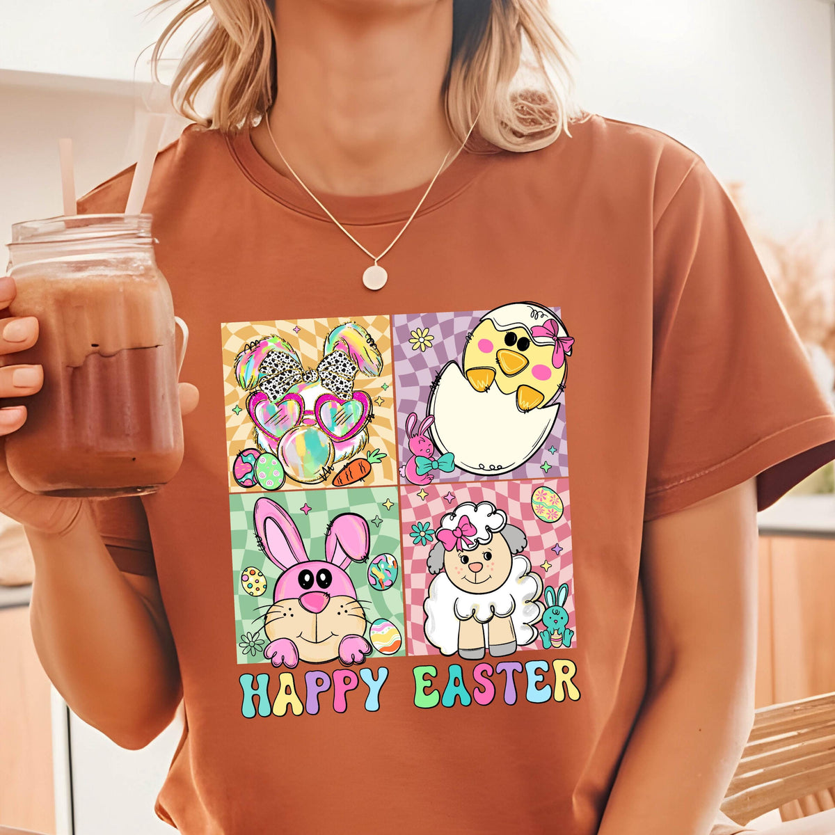 May include: A brown t-shirt with a graphic print of four cartoon animals in colorful squares. The animals are a bunny, a chick, a lamb, and a bunny wearing sunglasses. The text "Happy Easter" is printed below the squares.