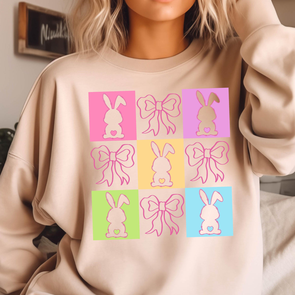 May include: A light beige sweatshirt with a colorful design of pink bunnies and bows. The design is in a grid pattern with each square having a different pastel color.