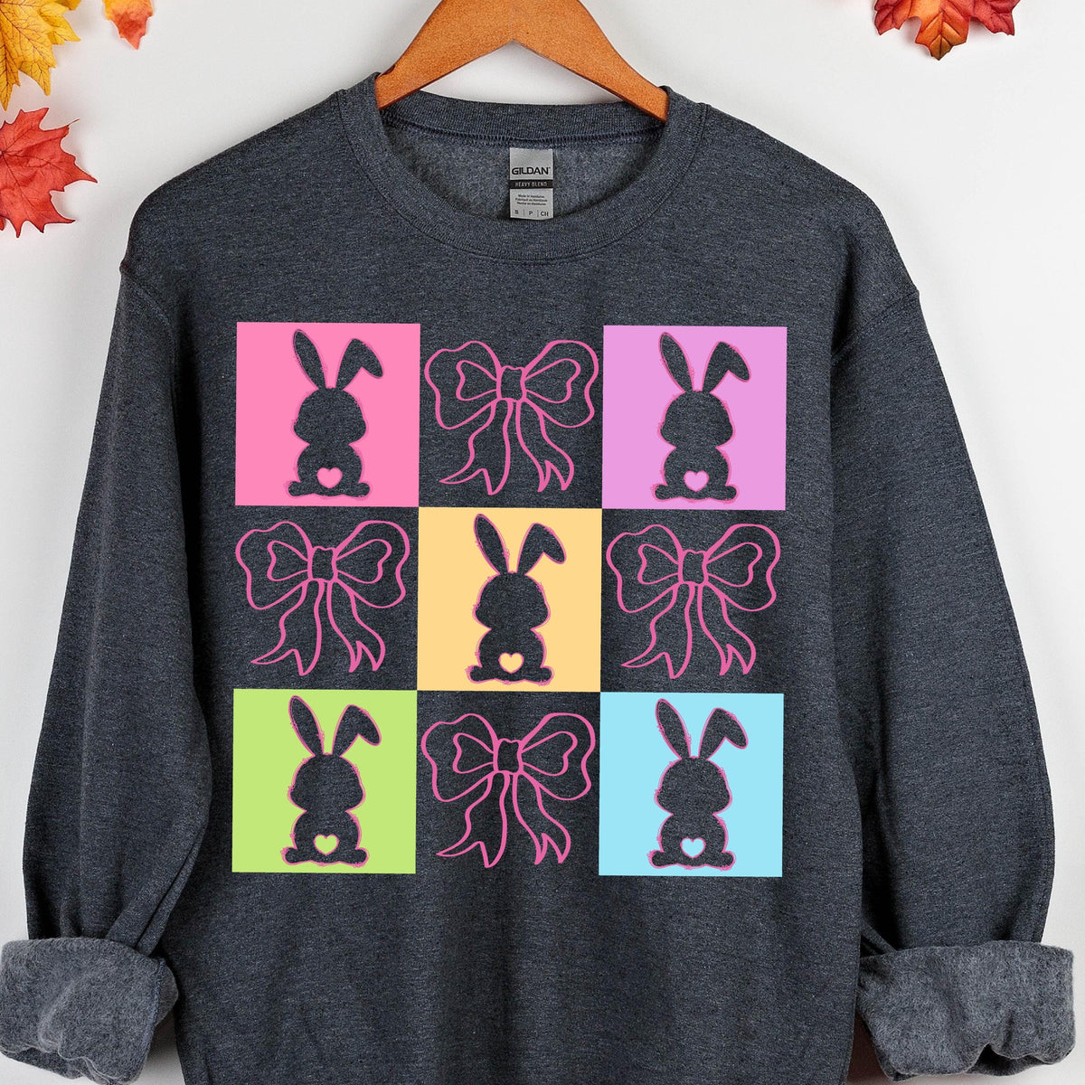 May include: A dark gray sweatshirt with a colorful pattern of nine squares. Each square features a silhouette of a bunny rabbit with a heart and a bow. The squares are in alternating colors of pink, yellow, light blue, and green.