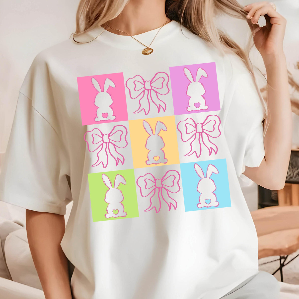 May include: White sweatshirt featuring a grid of colorful squares with Easter-themed designs. Each square contains either a bunny silhouette with a heart or a bow. The colors include pink, purple, yellow, green, and blue.
