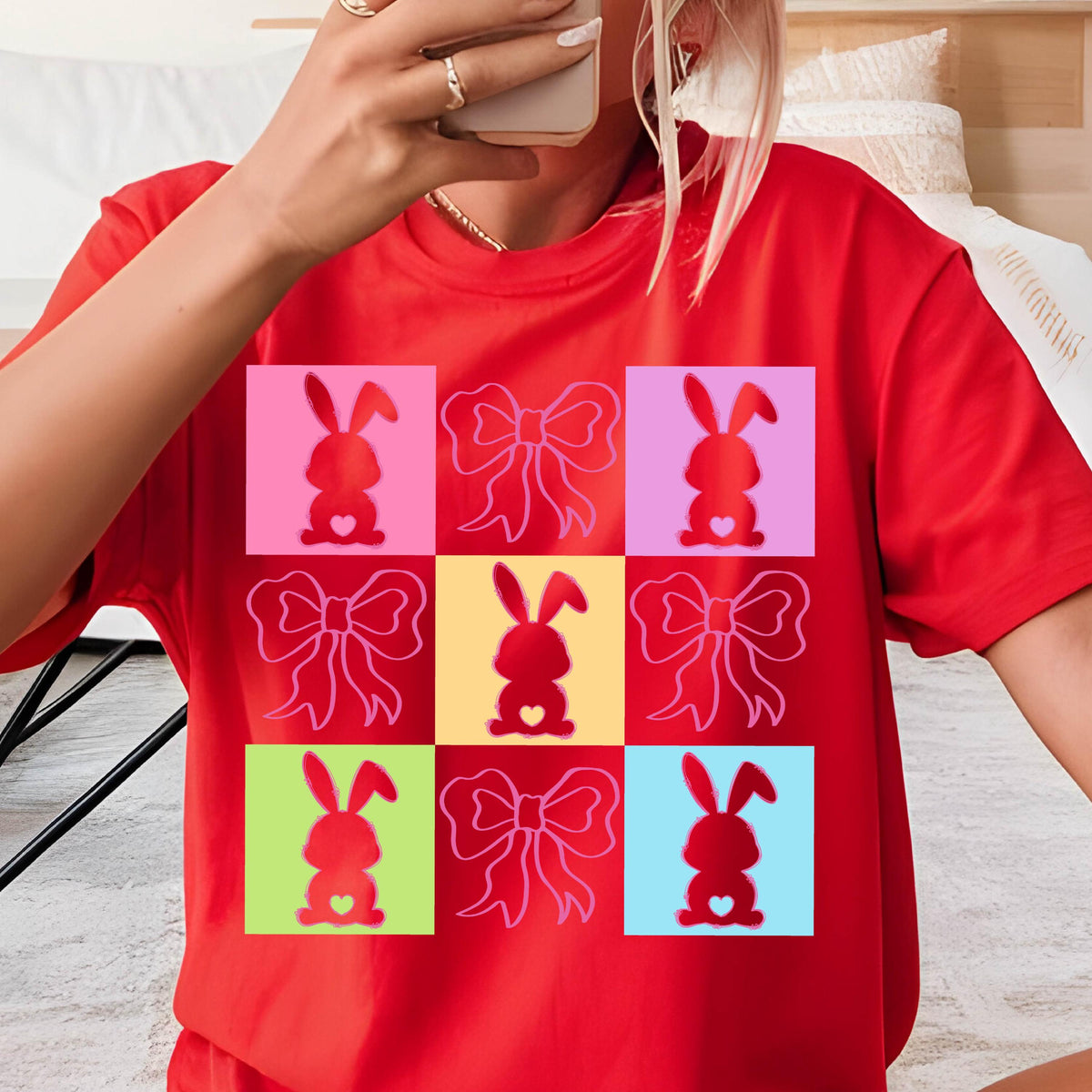 May include: Red t-shirt featuring a colorful grid pattern of bunny silhouettes and bows. The bunnies are red with heart-shaped bellies, set against pink, yellow, and blue squares. The bows are outlined in white.