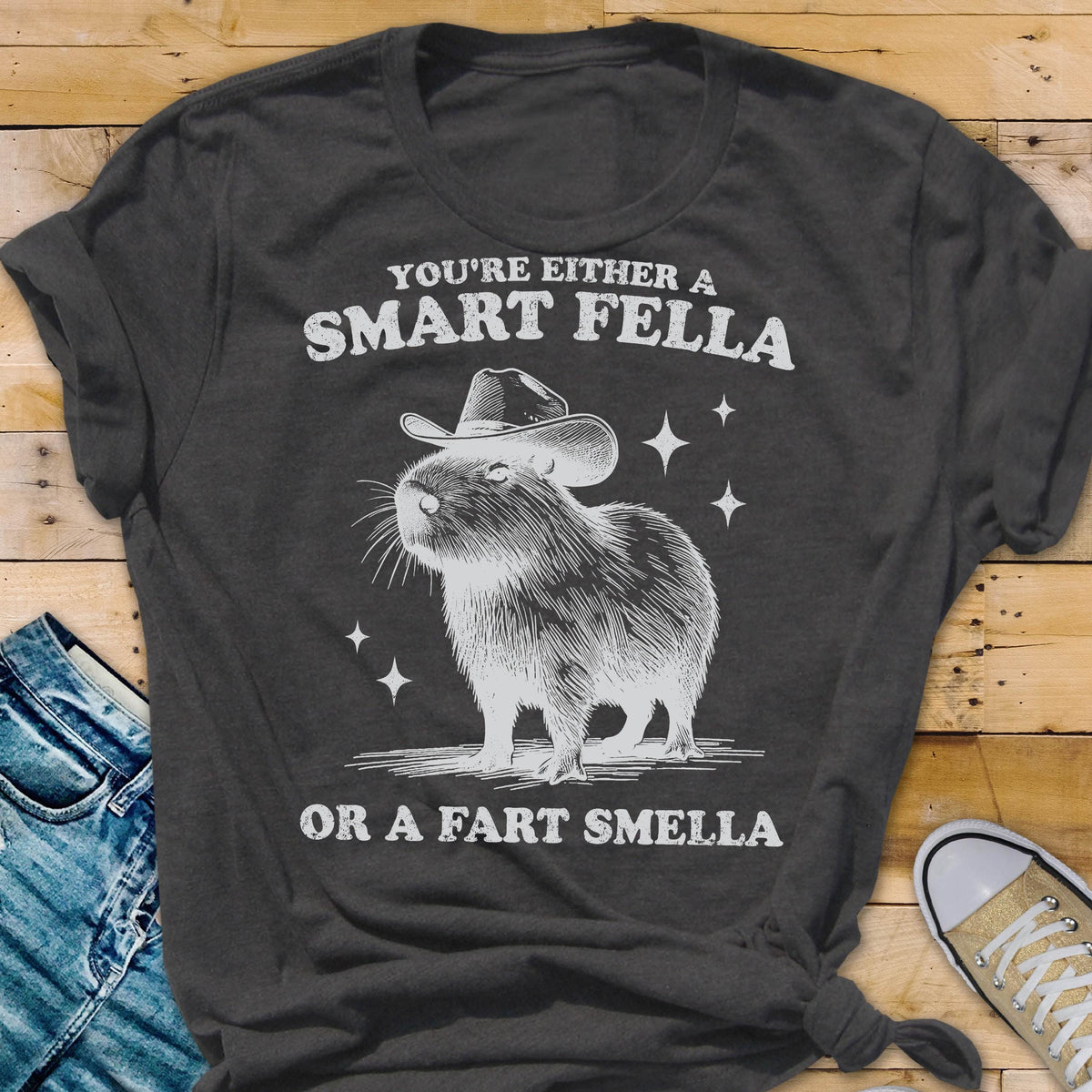 May include: Dark gray t-shirt with a graphic of a capybara wearing a cowboy hat. The text reads "YOU'RE EITHER A SMART FELLA OR A FART SMELLA" in white. The design includes star accents.