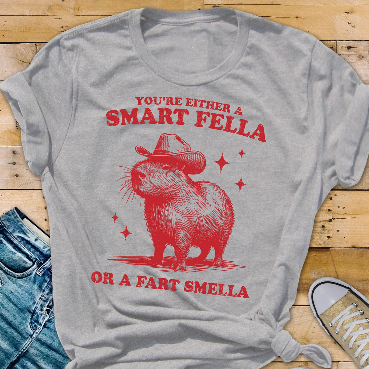 May include: A light gray t-shirt with a red graphic of a capybara wearing a cowboy hat. The text reads "YOU'RE EITHER A SMART FELLA OR A FART SMELLA" in red. The design includes red star accents.