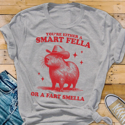 May include: A light gray t-shirt with a red graphic of a capybara wearing a cowboy hat. The text reads "YOU'RE EITHER A SMART FELLA OR A FART SMELLA" in red. The design includes red star accents.