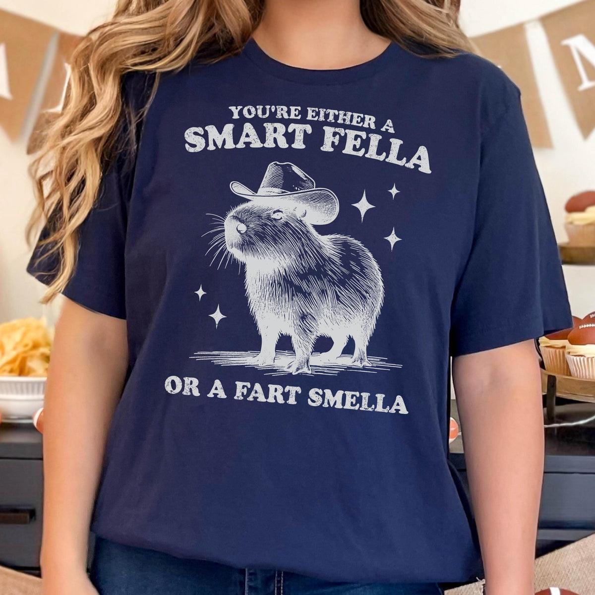 May include: Navy blue t-shirt with a graphic of a capybara wearing a cowboy hat and the text "YOU'RE EITHER A SMART FELLA OR A FART SMELLA" in white. The design includes star accents.