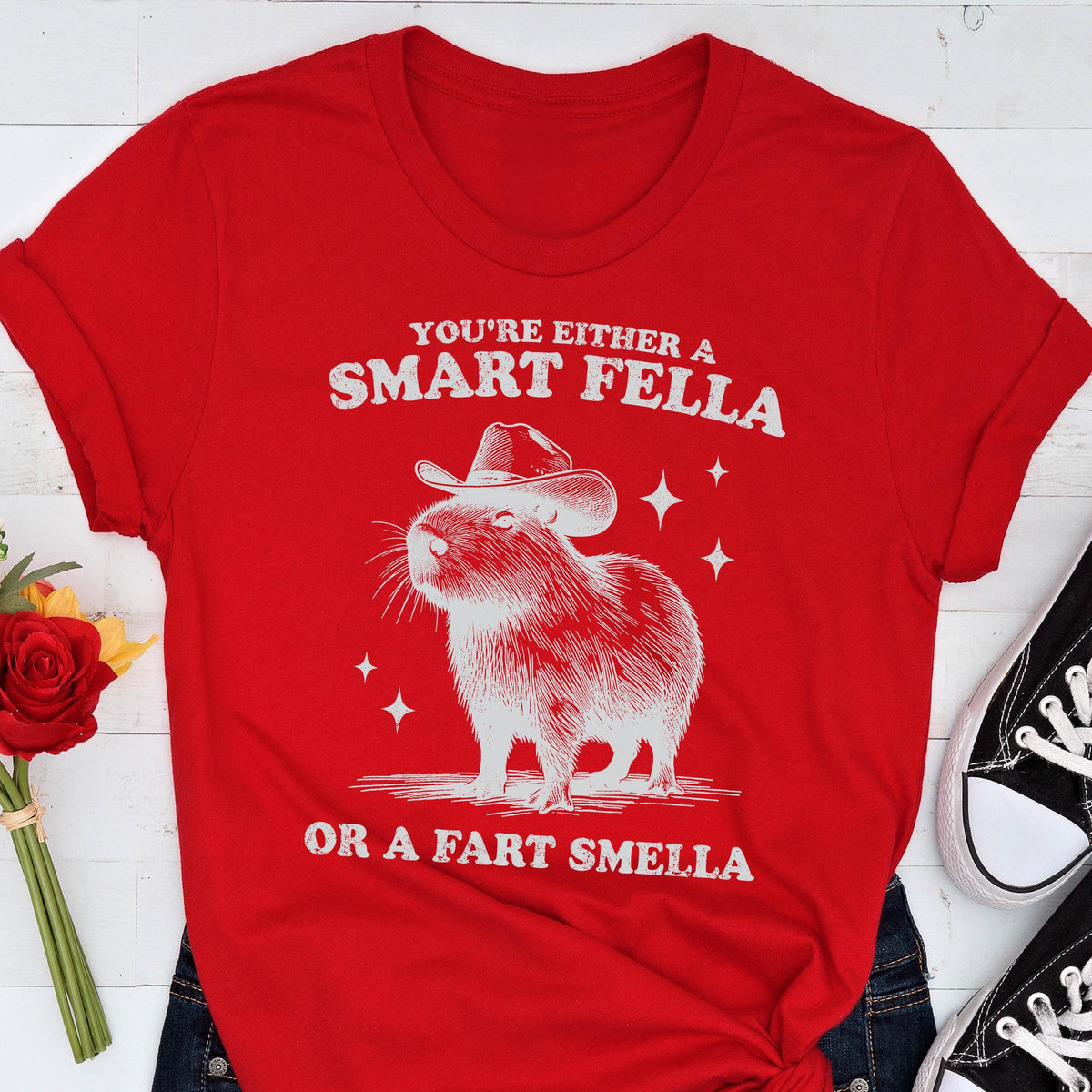 May include: Red t-shirt with a white graphic of a capybara wearing a cowboy hat. The text reads "YOU'RE EITHER A SMART FELLA OR A FART SMELLA" in white. The shirt is paired with blue jeans and black sneakers.