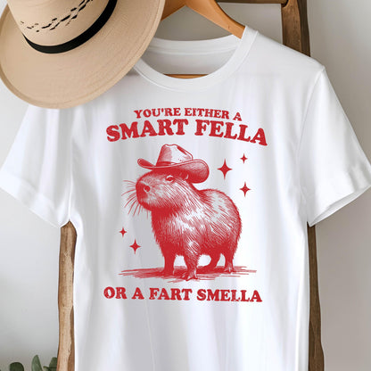 May include: White t-shirt with a red graphic of a capybara wearing a cowboy hat. The text reads "YOU'RE EITHER A SMART FELLA OR A FART SMELLA" in red. A tan cowboy hat hangs in the background.