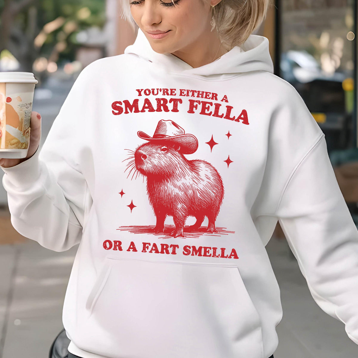 May include: White hoodie featuring a red illustration of a capybara wearing a cowboy hat. The text reads "YOU'RE EITHER A SMART FELLA OR A FART SMELLA" in red. Four red star shapes are also included in the design.