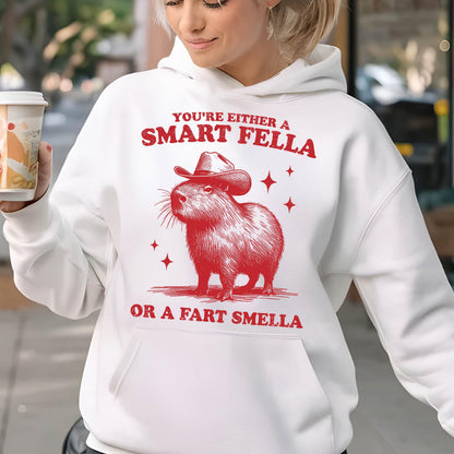 May include: White hoodie featuring a red illustration of a capybara wearing a cowboy hat. The text reads "YOU'RE EITHER A SMART FELLA OR A FART SMELLA" in red. Four red star shapes are also included in the design.