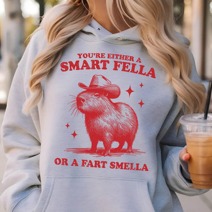 May include: A light gray hoodie featuring a red illustration of a capybara wearing a cowboy hat. The text above the capybara reads "YOU'RE EITHER A SMART FELLA" and below it reads "OR A FART SMELLA".