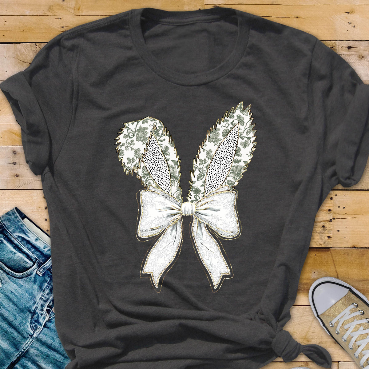 May include: A charcoal gray t-shirt with a white bow and two green bunny ears with gold accents. The bow and ears are a graphic print on the shirt.