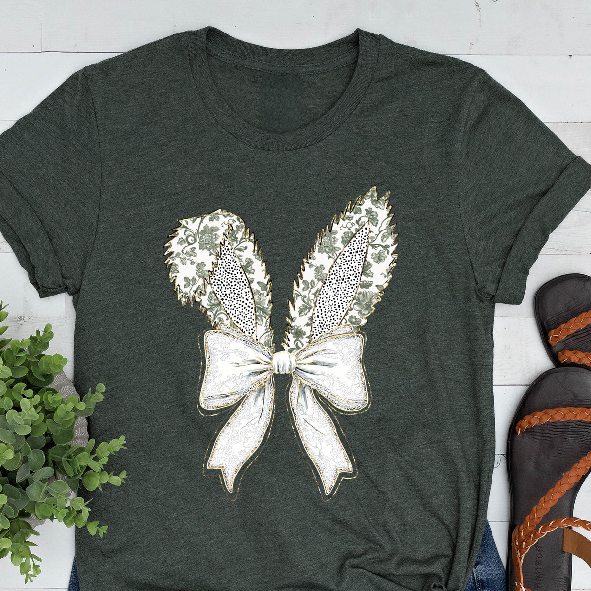 May include: A dark green t-shirt with a white bow and bunny ears design. The bow and ears are decorated with a floral pattern and gold glitter.