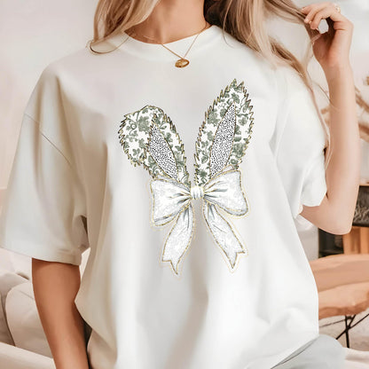 May include: White t-shirt with a gold and green bow design. The bow is made of a floral pattern and has a gold outline.