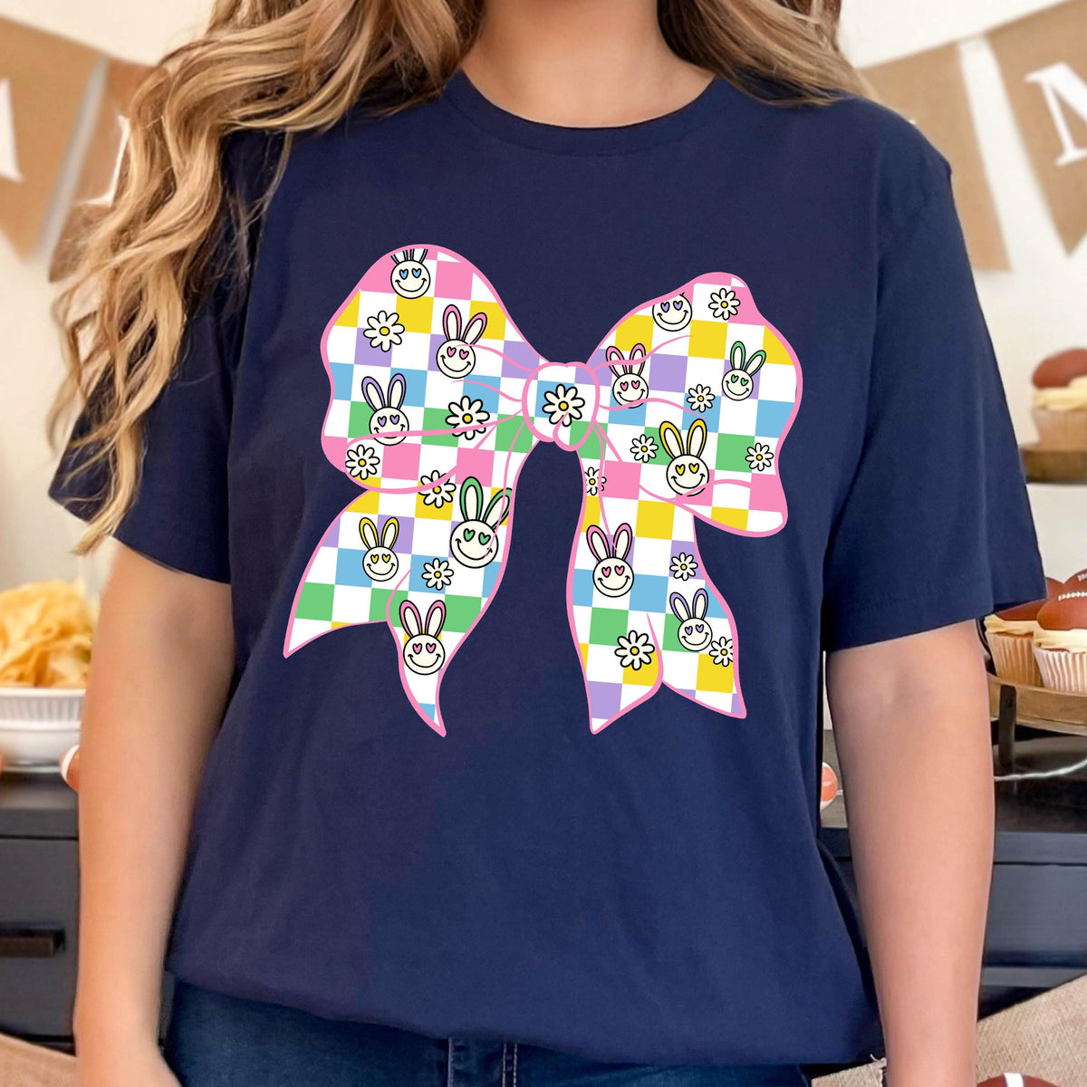 May include: A navy blue t-shirt with a pink bow printed on the front. The bow is checkered with yellow, green, blue, and purple squares. Each square has a small white flower or a cartoon bunny with a smiley face.