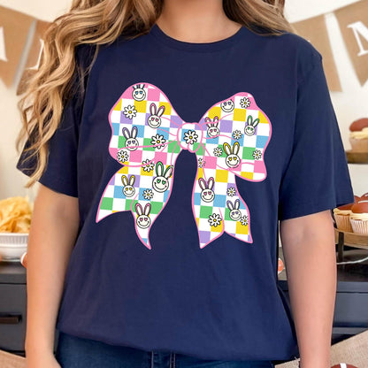 May include: A navy blue t-shirt with a pink bow printed on the front. The bow is checkered with yellow, green, blue, and purple squares. Each square has a small white flower or a cartoon bunny with a smiley face.