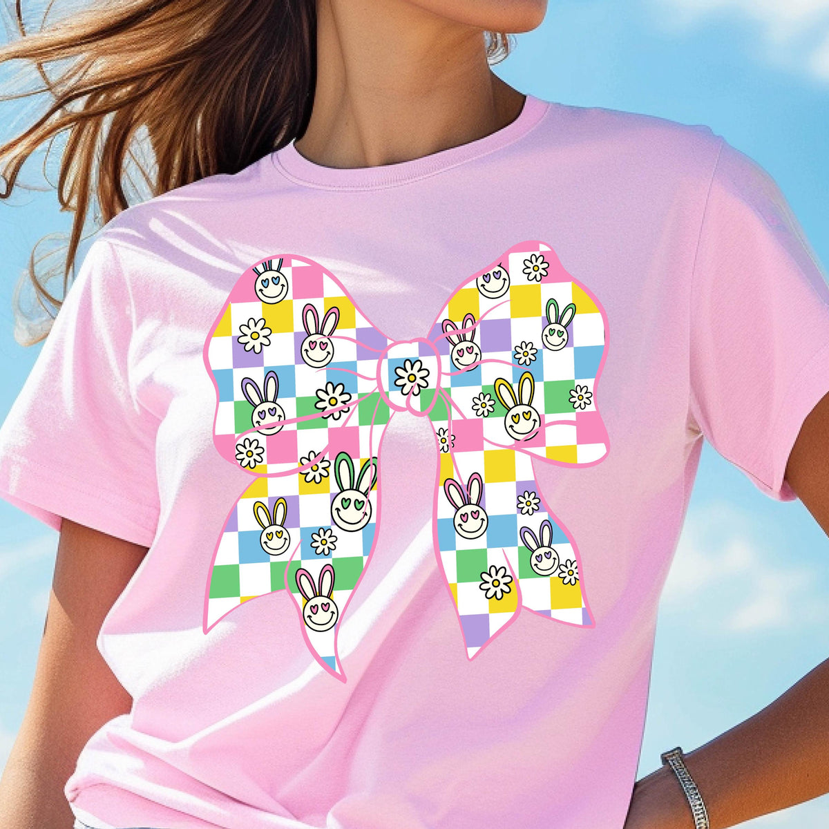 May include: A pink t-shirt with a large, colorful bow printed on the front. The bow is made up of checkered squares in shades of pink, purple, green, yellow, and blue. Each square has a small, white daisy and a cartoon bunny with a smiley face.